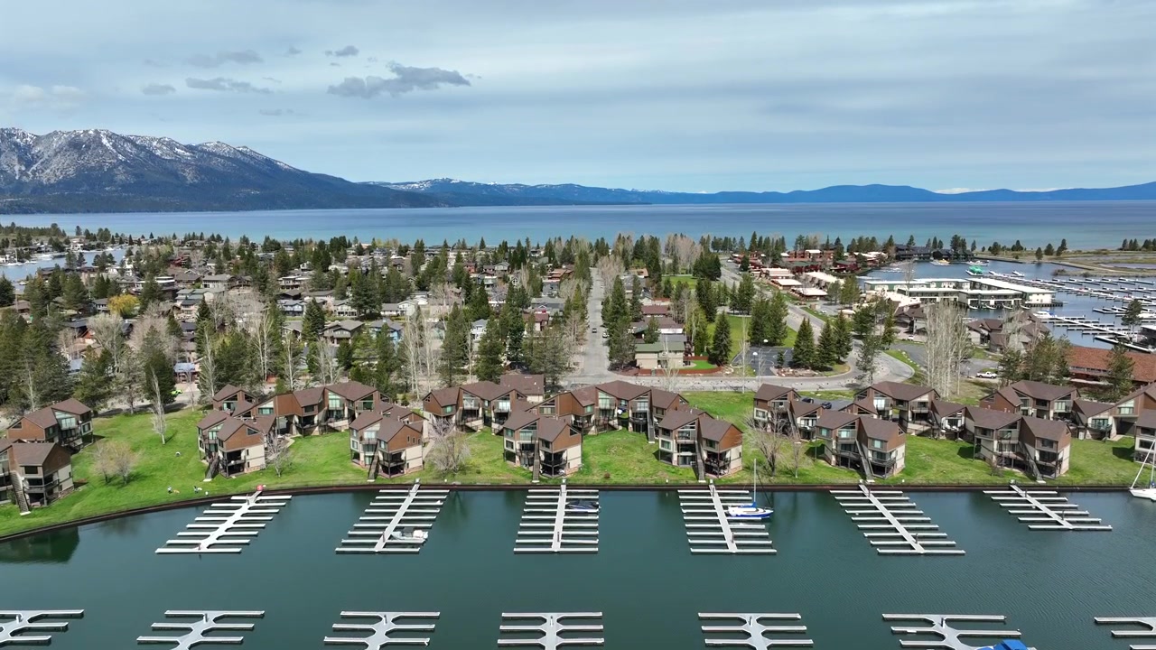 Marina di South Lake Tahoe con moli per barche vuoti e case sul lungomare sullo sfondo di foreste e montagne innevate sotto un cielo nuvoloso. Paesaggio lacustre panoramico in California, Stati Uniti. Ripresa aerea in salita.