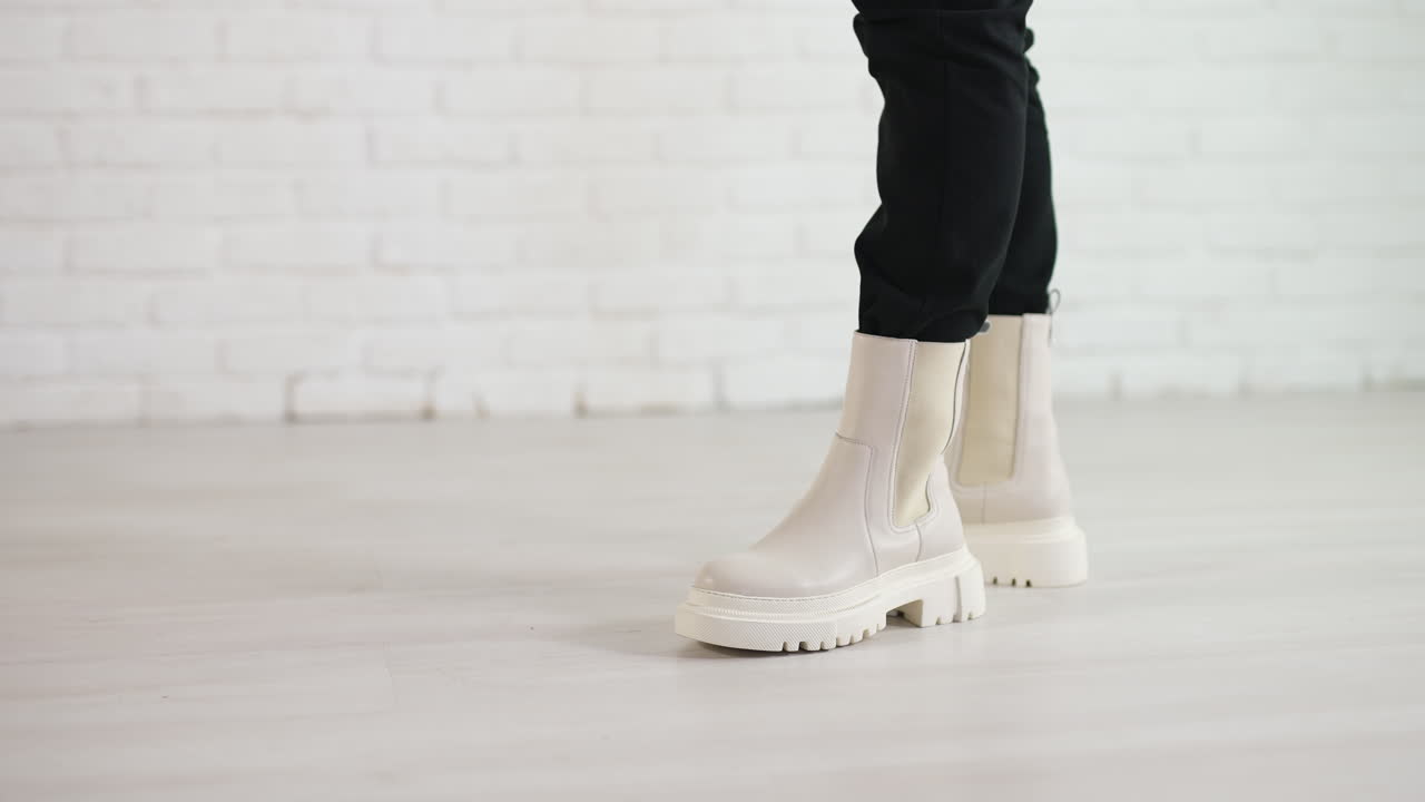 Woman in black jeans walks in new stylish white boots. Demonstrating the shoes of top fashion. White brick wall backdrop.
