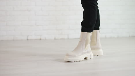 woman in black jeans walks in new stylish white boots. demonstrating the shoes of top fashion. white brick wall backdrop.