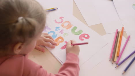 top view of blonde little girl drawing the phrase save the earth on a paper on a table in craft workshop