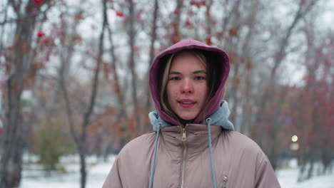 young lady smiling slightly at camera, wearing winter jacket with hood, outdoors, blurred background with trees and snow, casual winter portrait, cold weather, urban park setting