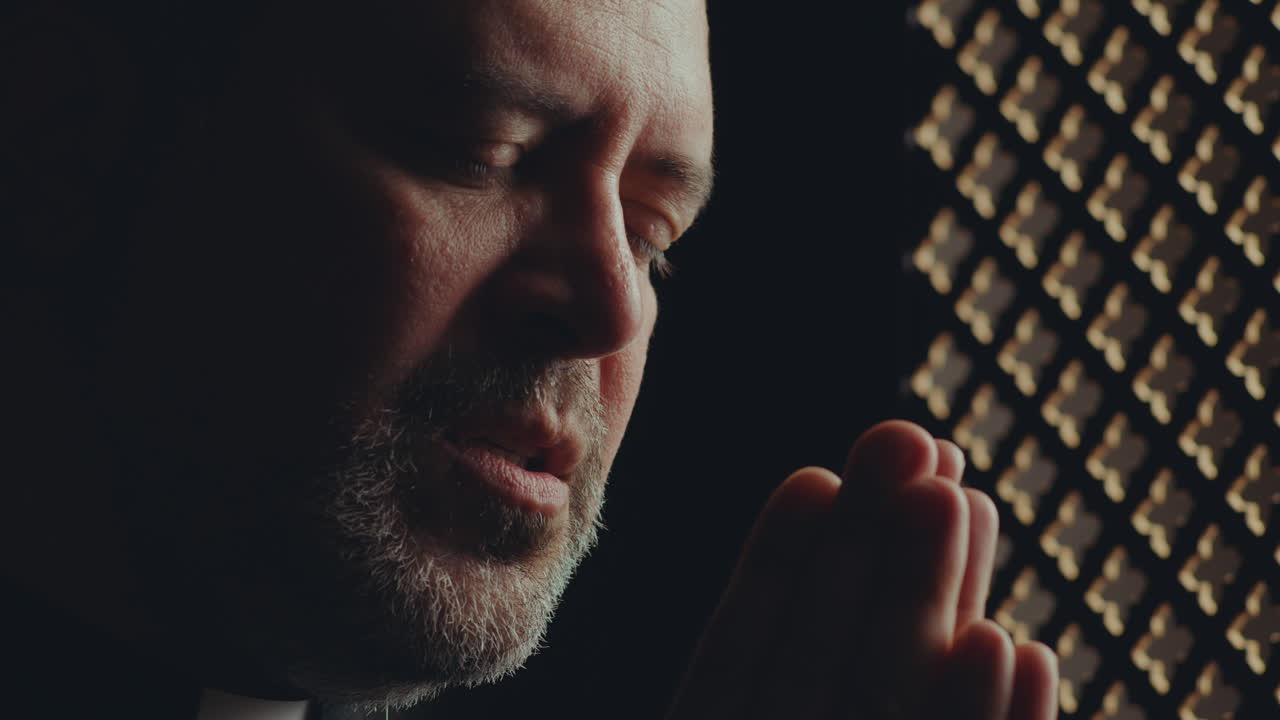 Priest Saying Prayer with Hands Clasped in Confessional Booth