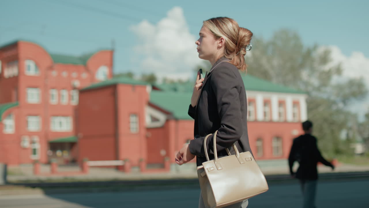 Side view of college student ending phone call, placing phone on chest while standing with handbag, passerby in front, urban setting with road, buildings, and cars in background