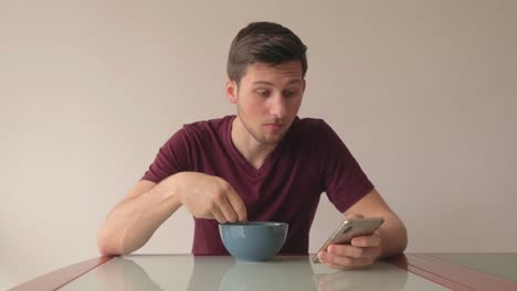 hombre sentado en la mesa comiendo cereales y reaccionando al contenido del teléfono móvil mientras se desplaza, aún filmado en interiores con luz natural