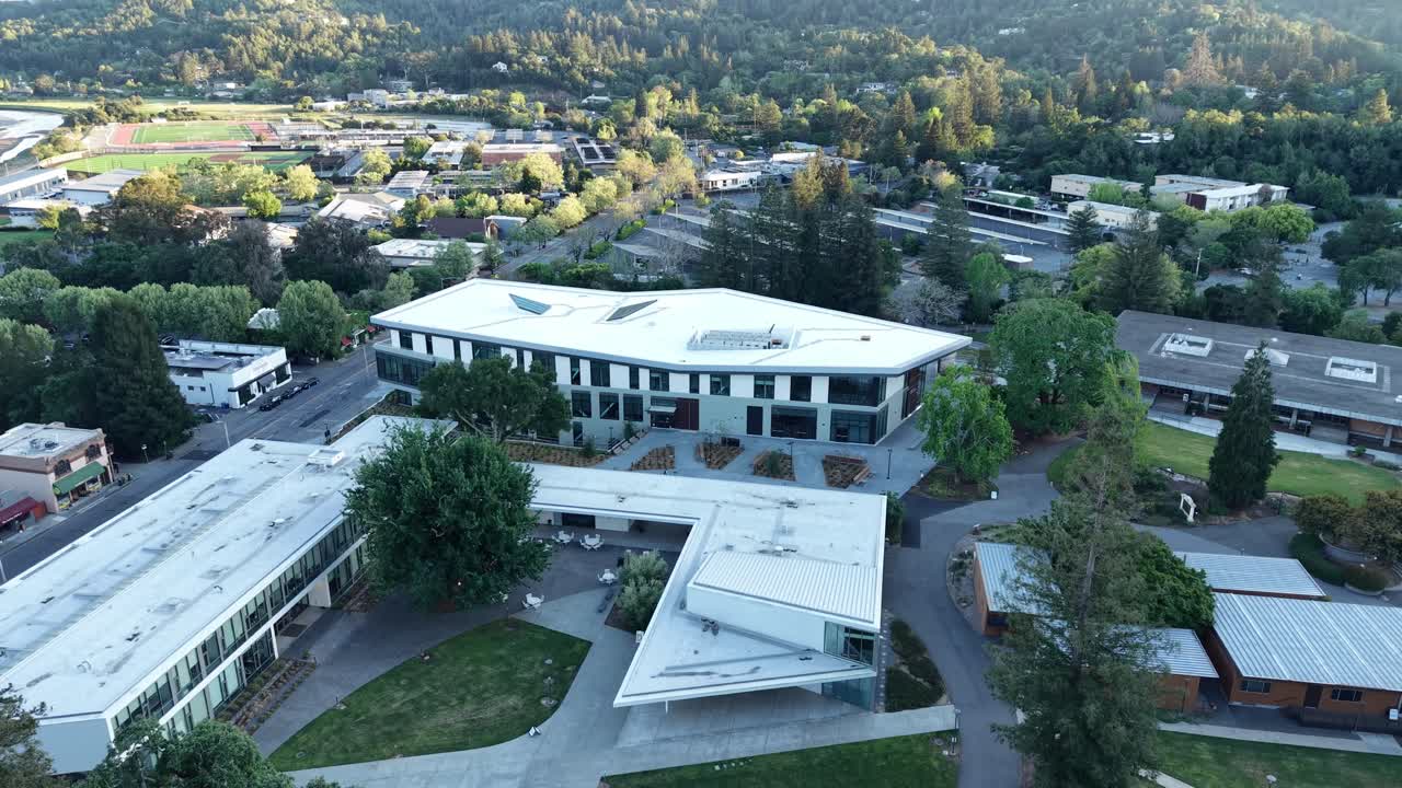 Tomada aérea de drone voando lentamente sobre a arquitetura moderna do campus do College of Marin em Kentfield, Califórnia, mostrando os edifícios acadêmicos cercados por árvores verdes exuberantes e colinas