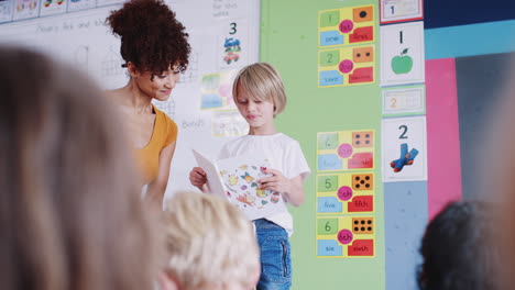 male pupil in elementary school classroom reading book to class with teacher