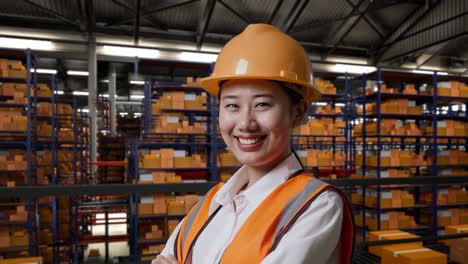 primer plano de una ingeniera asiática con casco de seguridad de pie en el almacén con estantes llenos de mercancías de entrega. cruzando los brazos y sonriendo a la cámara en el almacenamiento
