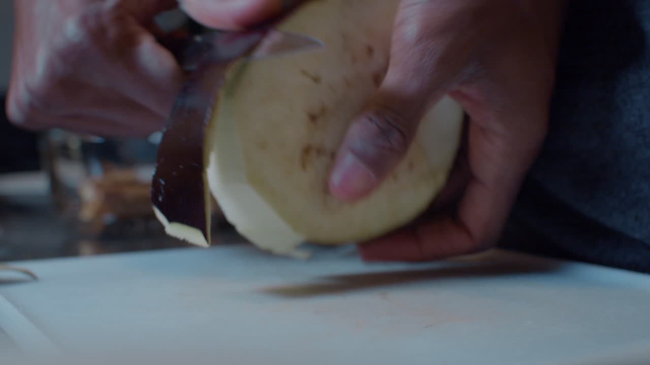 joven negro pelando media berenjena madura en una pizarra blanca de plástico en la cocina por la noche