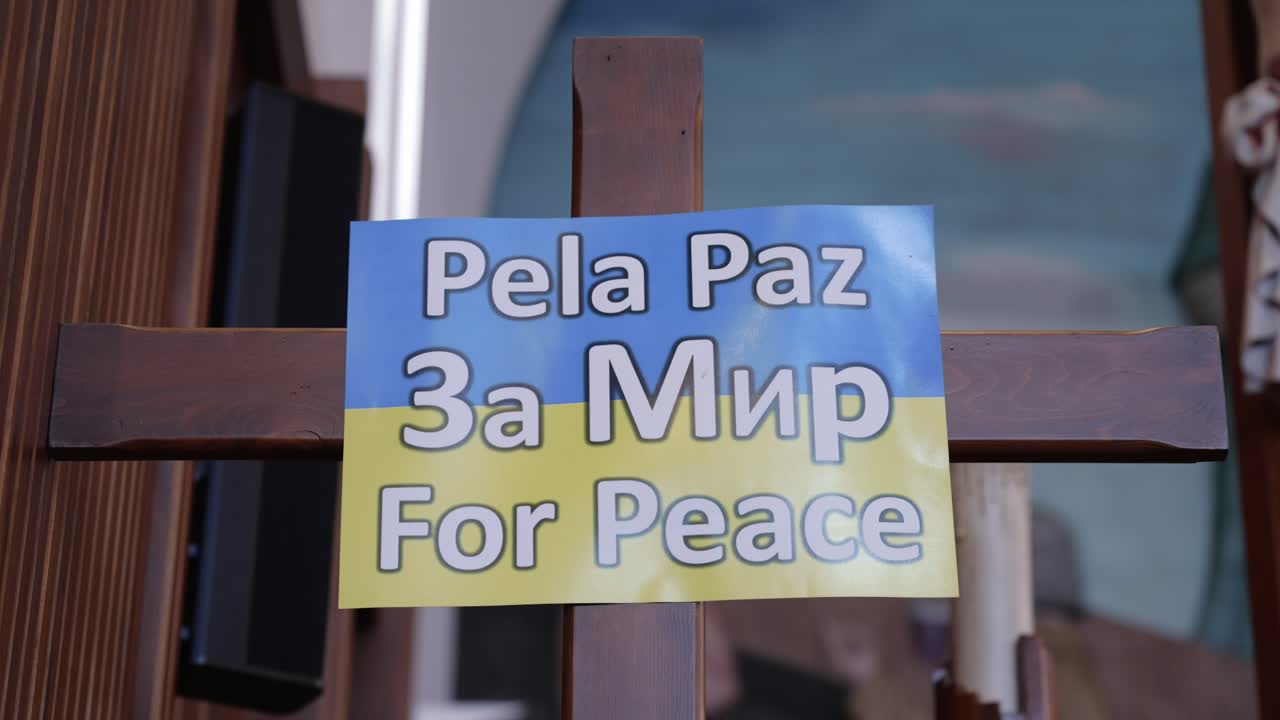 wooden cross inside a church with a blue and yellow sign reading "For Peace" in Portuguese, Russian, and English, supporting Ukraine