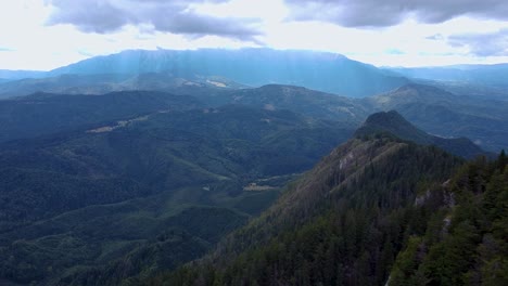 Vista-Lateral-De-Drones-De-Un-Exuberante-Bosque-Verde-De-Montaña-Con-Picos-Altos-En-El-Fondo-Rodeados-De-Rayos-Solares-Y-Nubes-Blancas-Esponjosas,-Ubicado-En-Poiana-Brasov,-Rumania