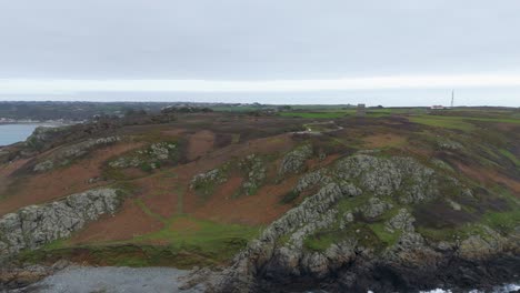 Guernsey-Pleinmont,-Vuelo-Desde-El-Mar-Hacia-El-Extremo-Suroeste-De-La-Isla-En-Un-Día-Nublado-Mostrando-Acantilados,-Brezales,-Rocas-De-Playa-Con-Olas-Espumosas,-Torre-De-Vigilancia-Alemana-Y-La-Torre-De-La-BBC