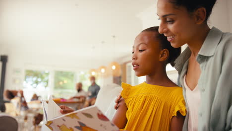 Madre-E-Hija-Leyendo-Un-Libro-En-El-Sofá-De-Casa-Con-Una-Familia-Multigeneracional-Detrás