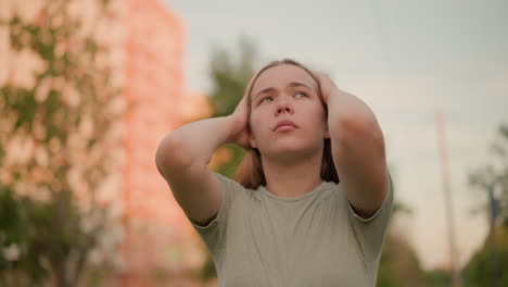 joven al aire libre con las manos presionadas contra su cabeza, los ojos cerrados, emanando una expresión contemplativa y de mal humor, rodeada de elementos urbanos y verdes borrosos