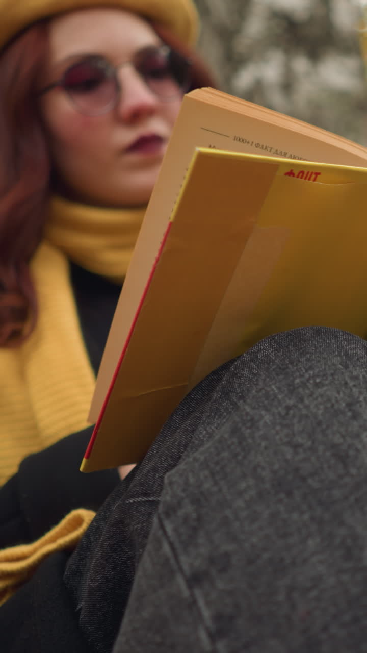 primer plano de un estudiante con gafas de sol, leyendo pensativo un libro cubierto de amarillo, las manos sujetan suavemente las páginas mientras el fondo presenta árboles de otoño borrosos