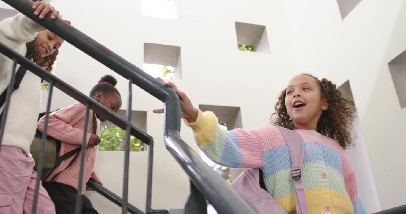African American girls heading downstairs after lead gesturing, holding rail, adjusting packs