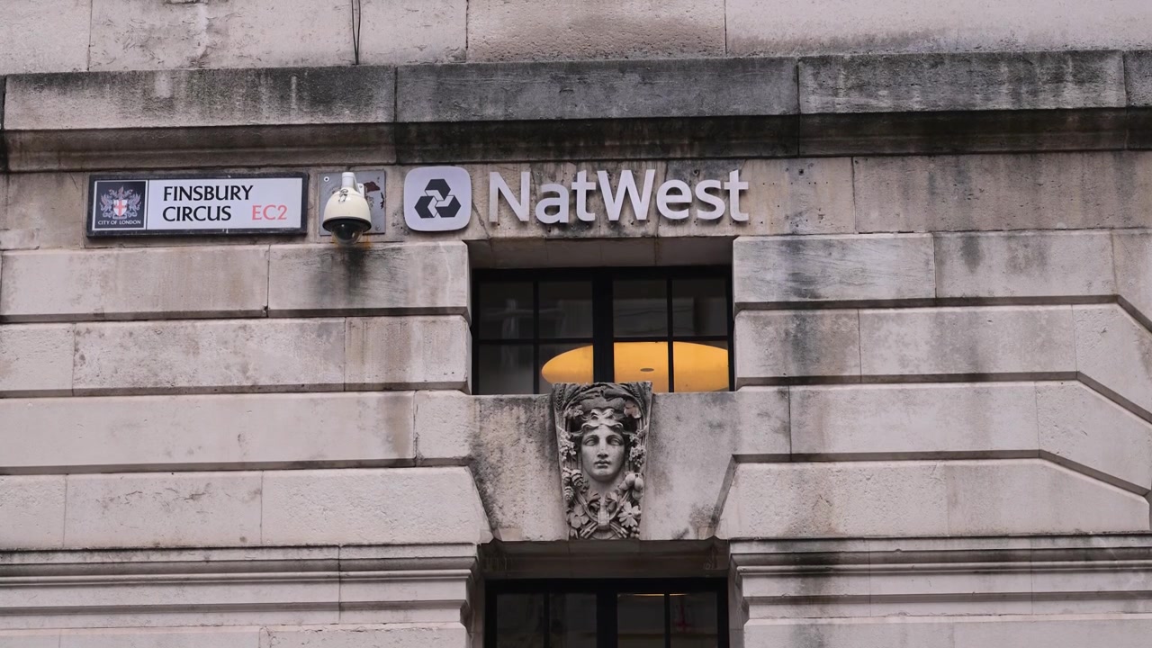 Il marchio NatWest, una telecamera a circuito chiuso e il cartello stradale Finsbury Circus EC2 appaiono su un muro di pietra in un'inquadratura statica ravvicinata