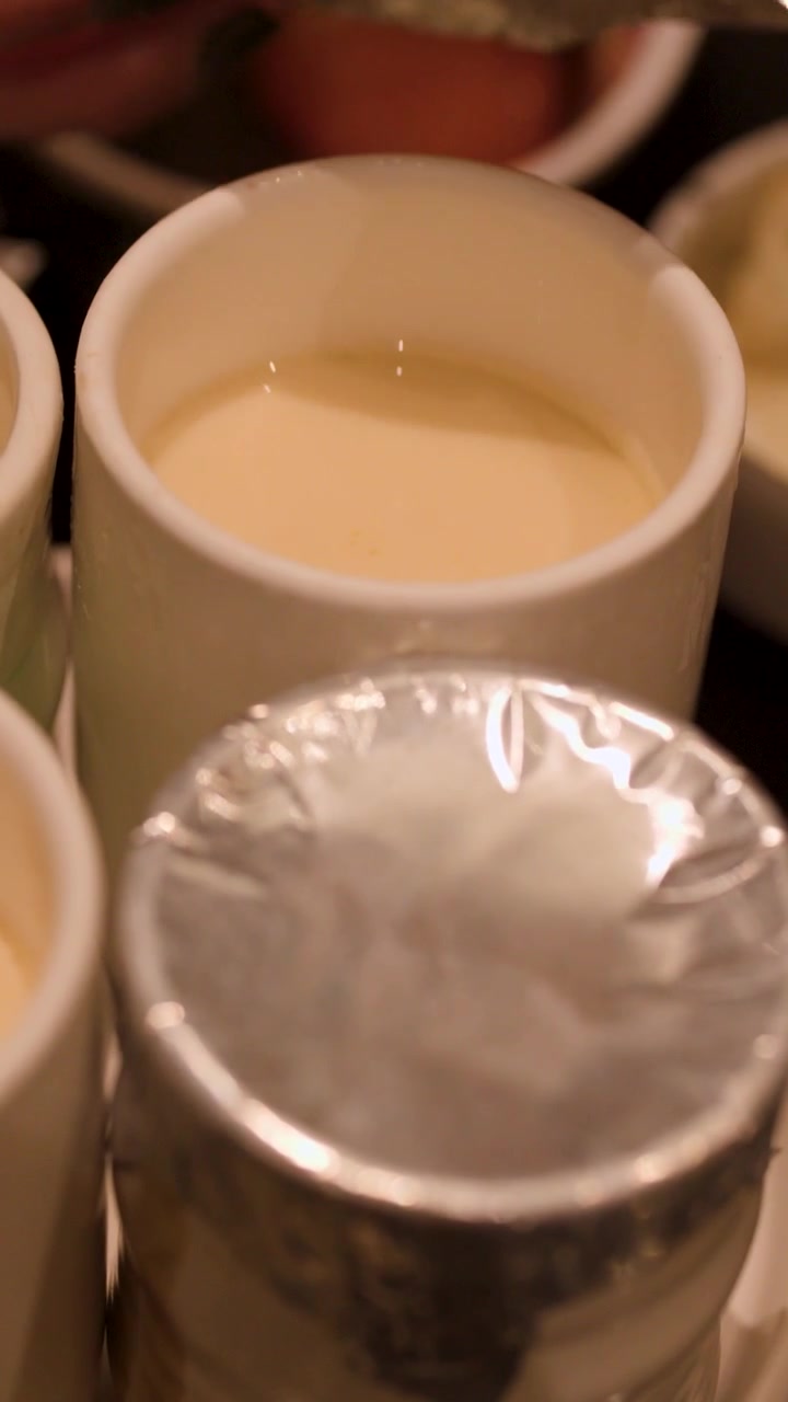 A close-up shot shows hands removing foil from cups of steamed egg custard in restaurant.