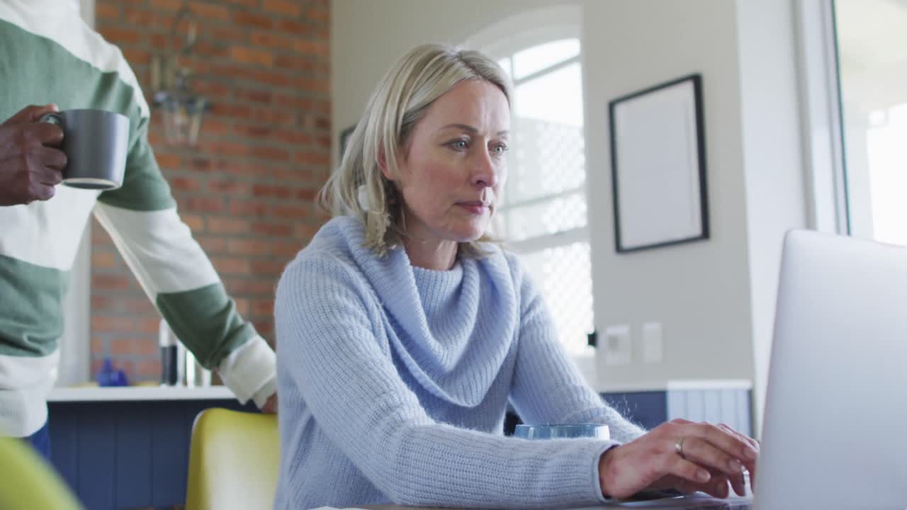 Stressed senior diverse couple in kitchen sitting at table, using laptop