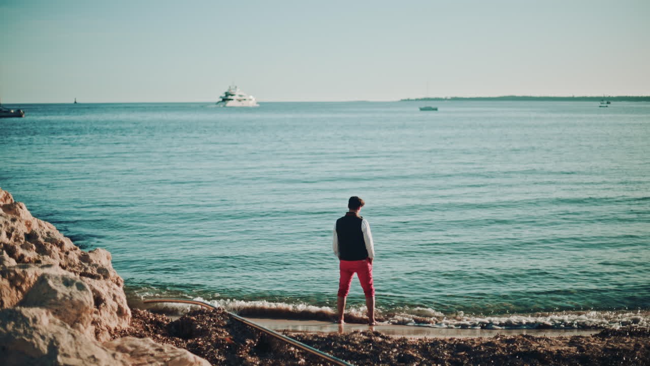 A man standing at the shoreline, facing the sea as small waves reach his feet