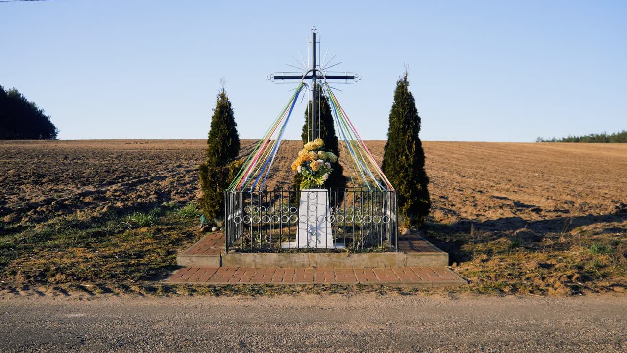 A sacred cross installation adorned with colorful ribbons and floral decorations in the countryside, surrounded by a serene natural environment during a sunny day, expressing spirituality and peace.