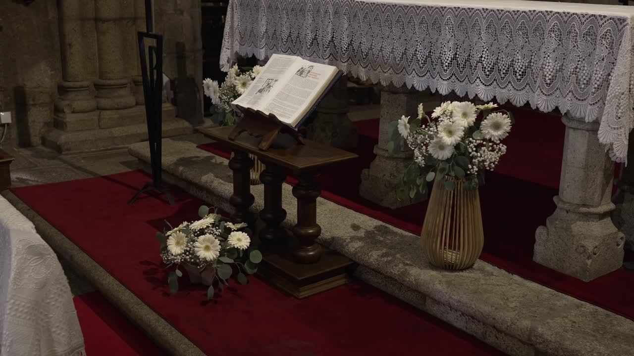 Open Bible on wooden stand by floral arrangements in a church altar with red carpet and stone base