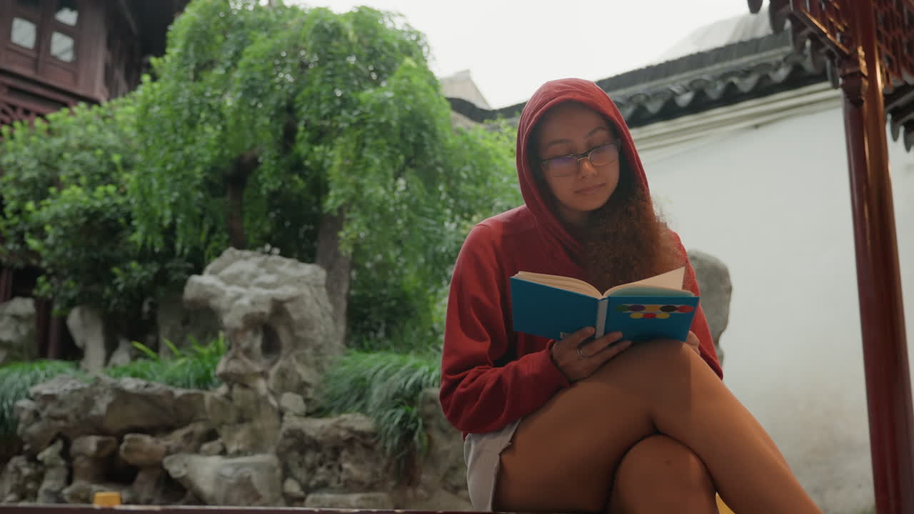 Genç Asyalı kadın, sakin, geleneksel bir avluda, ahşap bir pavyonun altında, taş kaya bahçesi ve söğüt ağacının yanında, kırmızı kapüşonlu sweatshirt giymiş, rahat bir duruşla kitap okurken ve günlüğünü incelerken, doğal ışık altında.