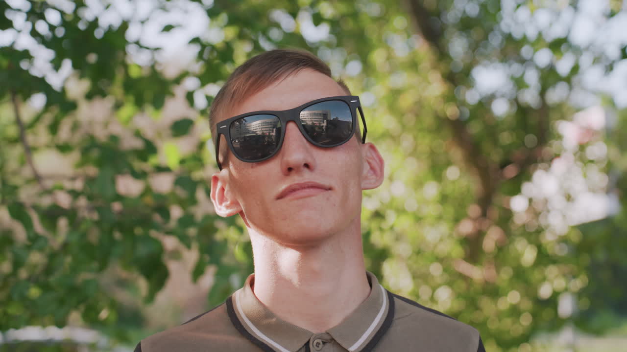 Young White Man Removing Sunglasses And Facing Camera With Confident, Composed Expression Leafy Green Backdrop, Soft Dappled Light, Short Hair, Casual Polo, Subtle Cinematic Framing, Mood Of Self