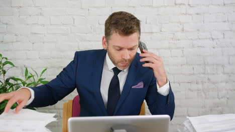 An-angry-businessman-is-becoming-frustrated-whilst-talking-on-the-phone-with-piles-of-paperword-in-his-office