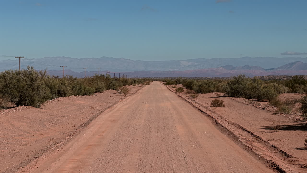 Drone a bassa quota che avanza su una strada sterrata isolata in un paesaggio desertico con montagne sullo sfondo e cielo azzurro limpido