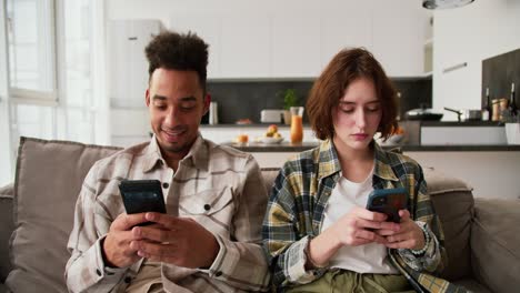 Calm-couple-a-cheerful-man-a-young-brunette-with-Black-skin-color-in-a-cream-plaid-shirt-scrolling-social-networks-while-sitting-on-the-sofa-with-his-girlfriend-who-is-also-looking-at-something-on-her-smartphone-in-a-modern-studio-apartment