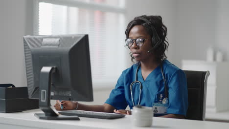 médica general negra afroamericana con bata blanca sentada en el escritorio de la oficina del médico y desplazando el ratón de la computadora mientras lee el historial médico del paciente