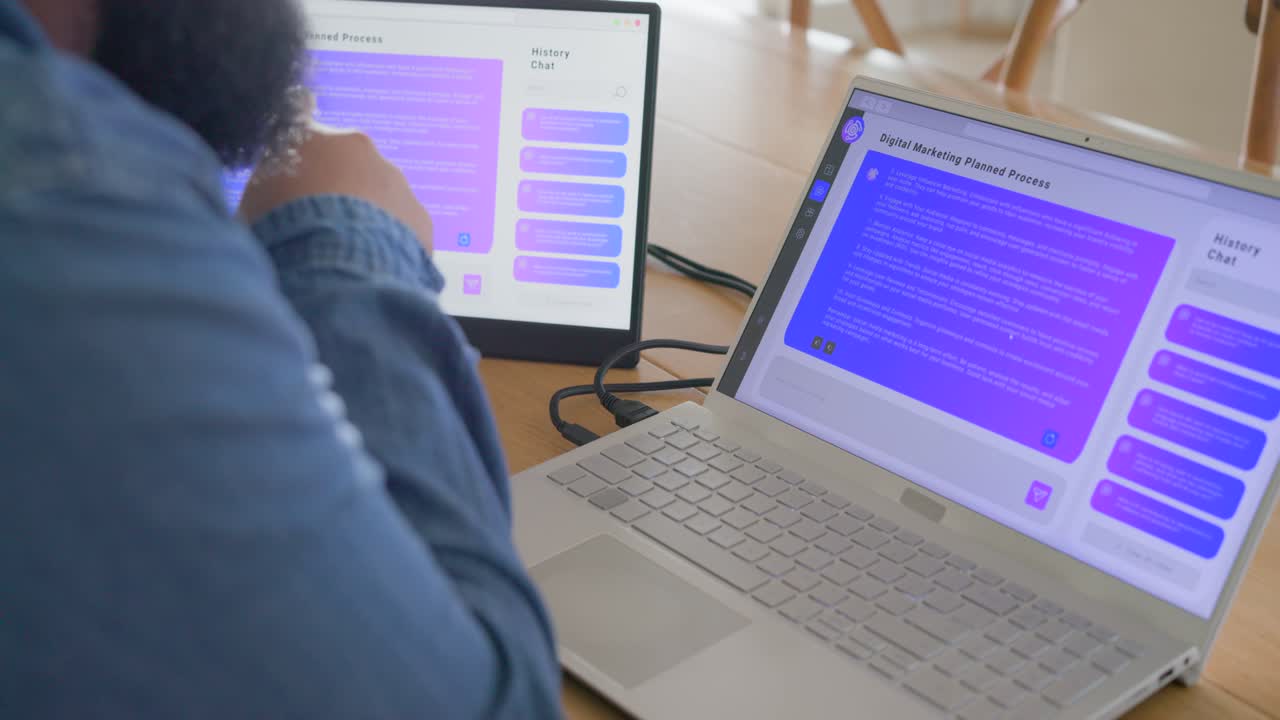 Adult African man reviewing purple chat on two-screen desk, tapping monitor and scrolling laptop