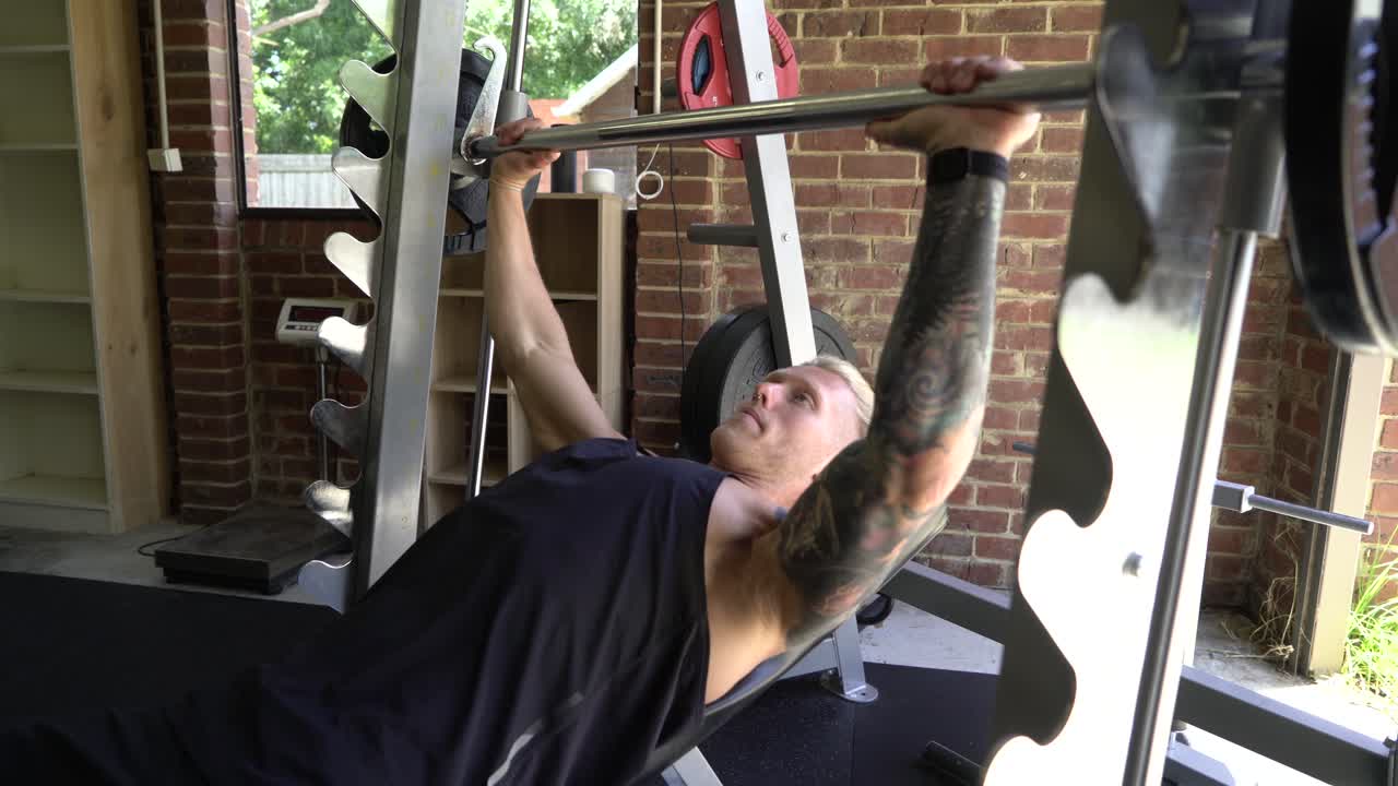 hombre tatuado en el gimnasio en casa haciendo press de banca inclinado con barra smith machine