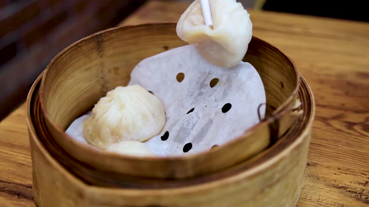 Chopsticks carefully pick up a soup dumpling from a bamboo steamer basket on a wooden table, under soft natural lighting with a close-up perspective