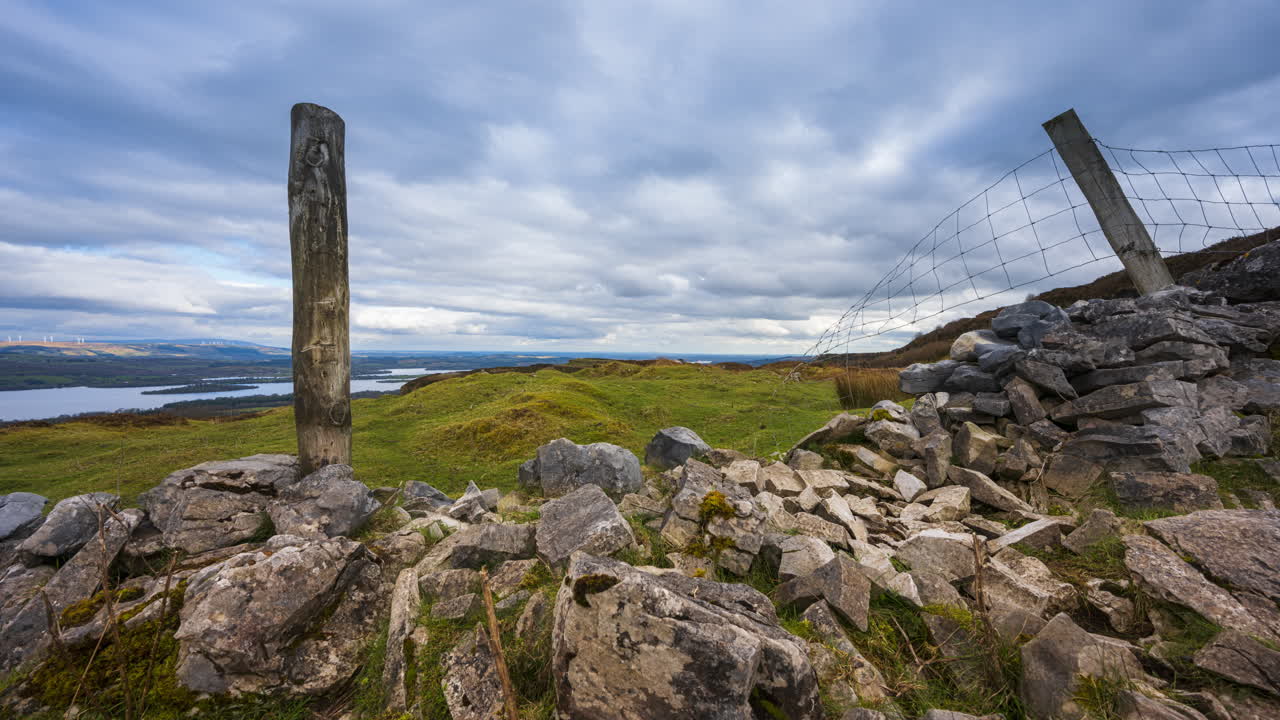 panorama motion timelapse de paisaje rural con pared de piedra, pilares de madera y ovejas en el campo de hierba y colinas y lago en la distancia durante un día nublado visto desde carrowkeel en el condado de sligo en irlanda