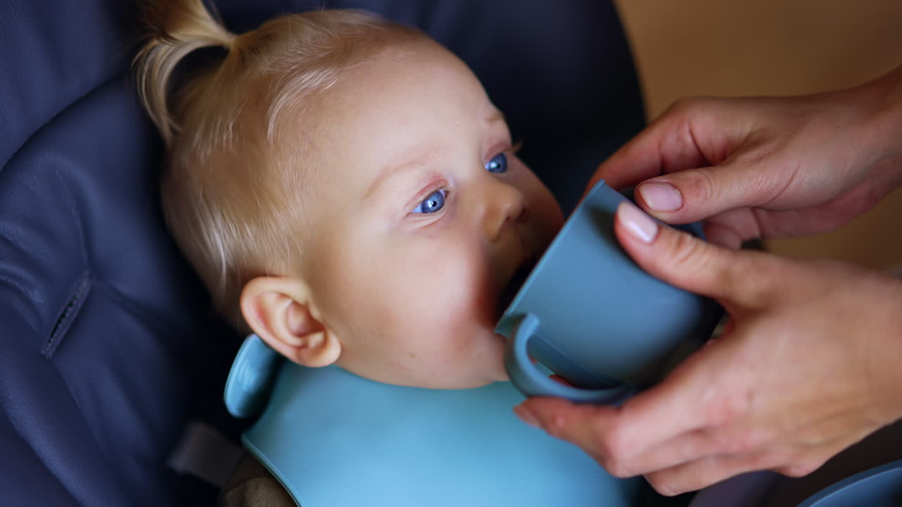 Cute Caucasian infant is given a cup to drink from. Close up. Mother is feeding her child.