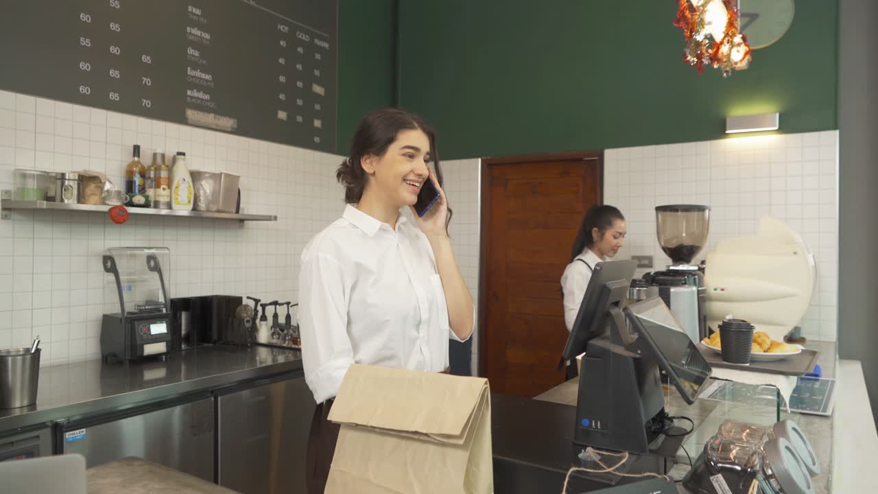 ocupada mujer barista de oriente medio gente en delantal recogiendo un pedido, hablando con el cliente por teléfono en el mostrador, cafetería caja. servicio de negocios. estilo de vida de la gente.
