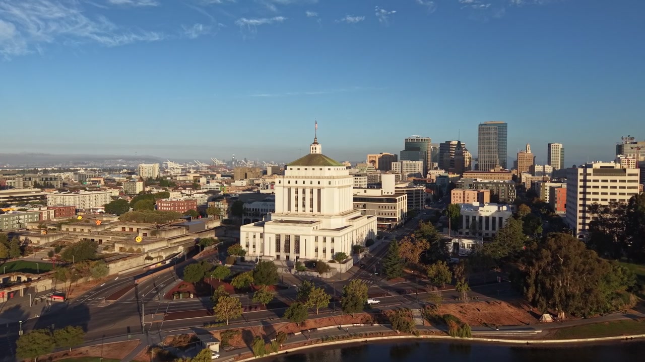 Luce dell'ora d'oro che illumina lo storico tribunale della contea di Alameda e il moderno skyline del centro di Oakland, con le acque del Lago Merritt visibili in primo piano. Ripresa con drone in avvicinamento.