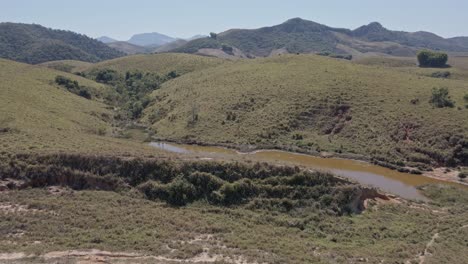 Subiendo-Detrás-Del-Pasto-En-La-Colina-Se-Ve-Una-Pequeña-Laguna-Con-Agua-Fangosa-Como-Un-Pantano-Escondido-Con-Montañas-En-El-Horizonte