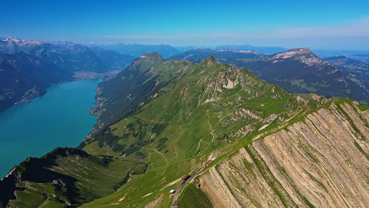una impresionante vista aérea de una cordillera en los alpes suizos con hierba verde, mirando sobre el pico más alto, con montañas cubiertas de nieve en el fondo y un lago azul brillante debajo