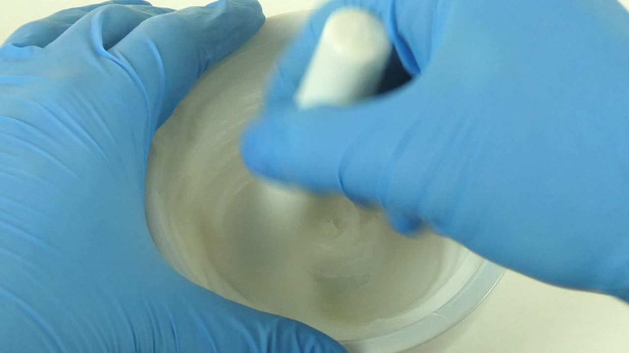 Macro shot of a cosmetic laboratory process where gloved hands in blue gloves mix a cream using a porcelain mortar and pestle.