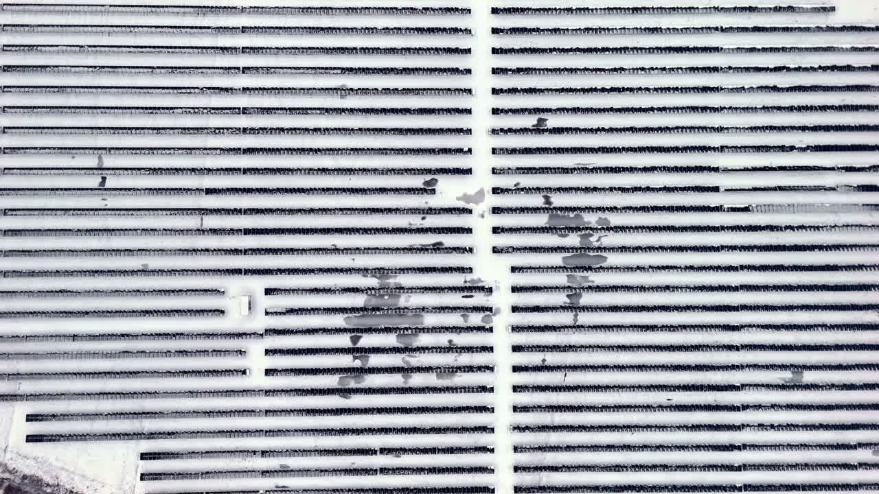 Top-down drone view of a snow-covered solar panel array in neat parallel rows, creating an abstract winter geometry of renewable power infrastructure