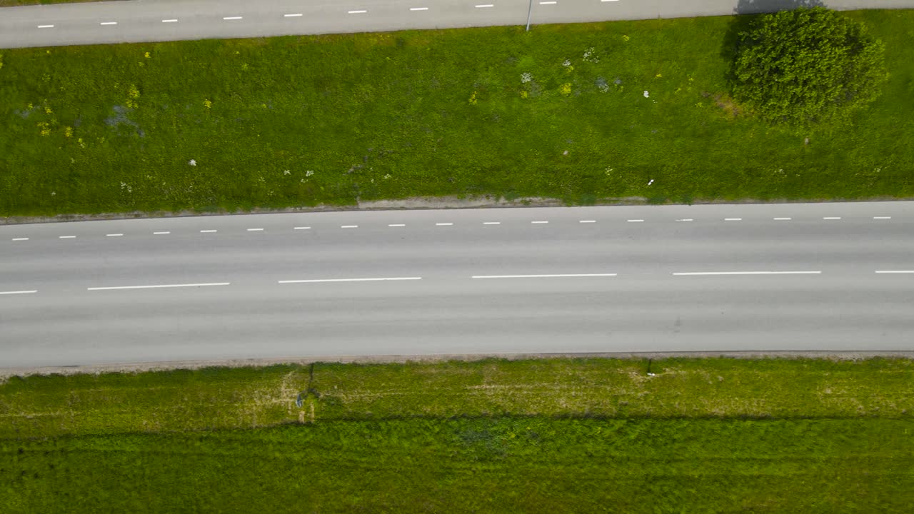 Top down aerial view of a rural and countryside paved or asphalt road where cars, vehicles and buses drive on back and fourth on a sunny summer day with green grass strip separating pedestrian road.