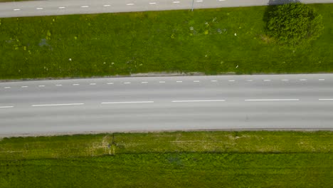 top down aerial view of a rural and countryside paved or asphalt road where cars, vehicles and buses drive on back and fourth on a sunny summer day with green grass strip separating pedestrian road.