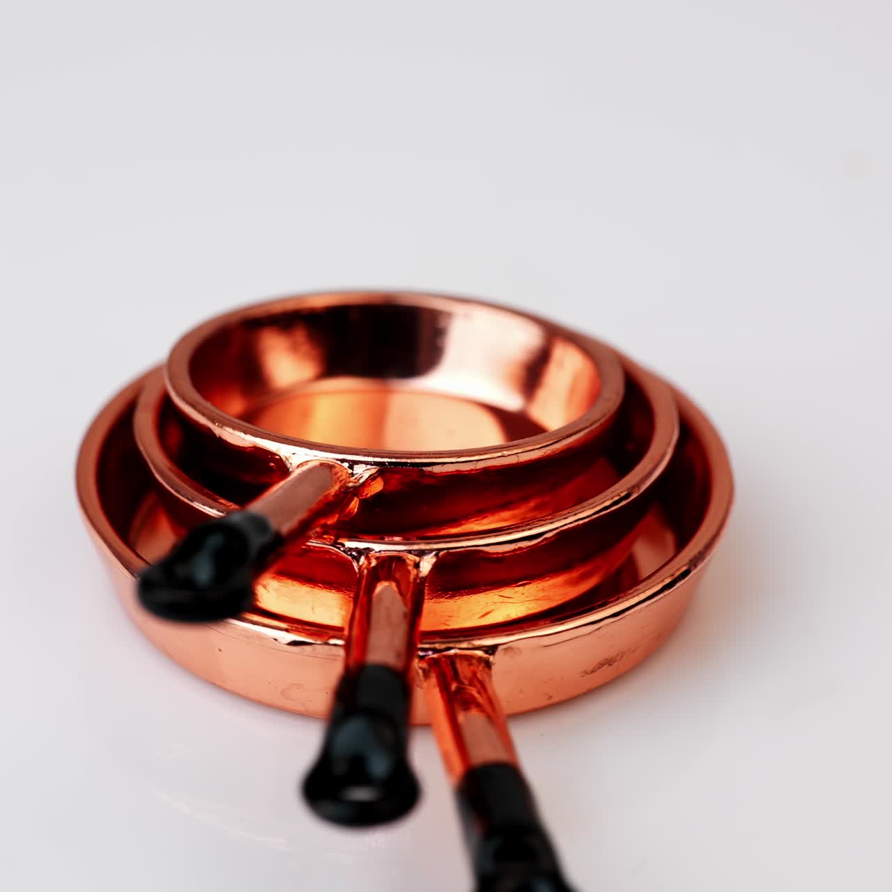A set of three copper frying pans with black handles piled on each other. Miniature crockery at rotating white background