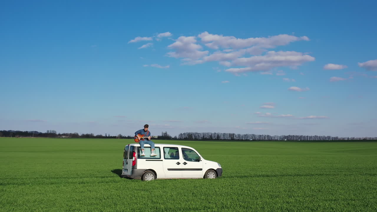 Young man with a guitar on a car moving on green field. Hipster guy likes extreme. Man playing the guitar while sitting on the top of a white car driving slowly in nature.