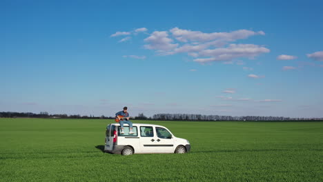 young man with a guitar on a car moving on green field. hipster guy likes extreme. man playing the guitar while sitting on the top of a white car driving slowly in nature.