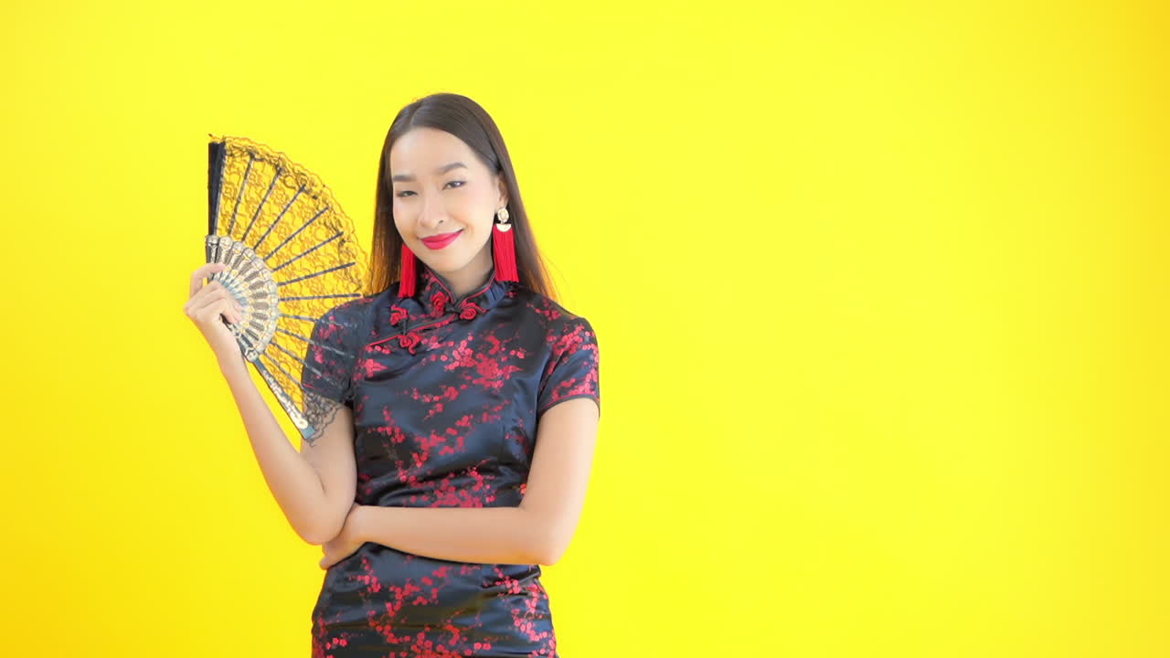 Asian female model in black and red silk dress opens black lace hand fan. Yellow background, copy space right. Attractive Chinese woman using ornate fan to cool self while smiling