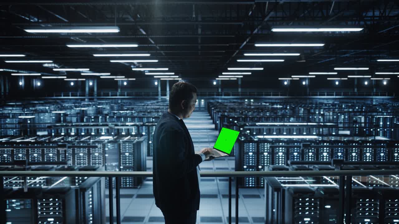 Portrait of IT Specialist Using Chroma Key Laptop in Data Center. Big Server Farm Cloud Computing Facility with Male Maintenance Administrator Working with Green Screen Computer. Medium Wide Arc Shot