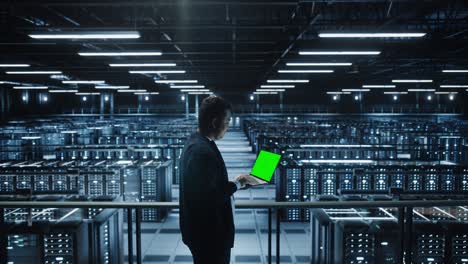 portrait of it specialist using chroma key laptop in data center. big server farm cloud computing facility with male maintenance administrator working with green screen computer. medium wide arc shot